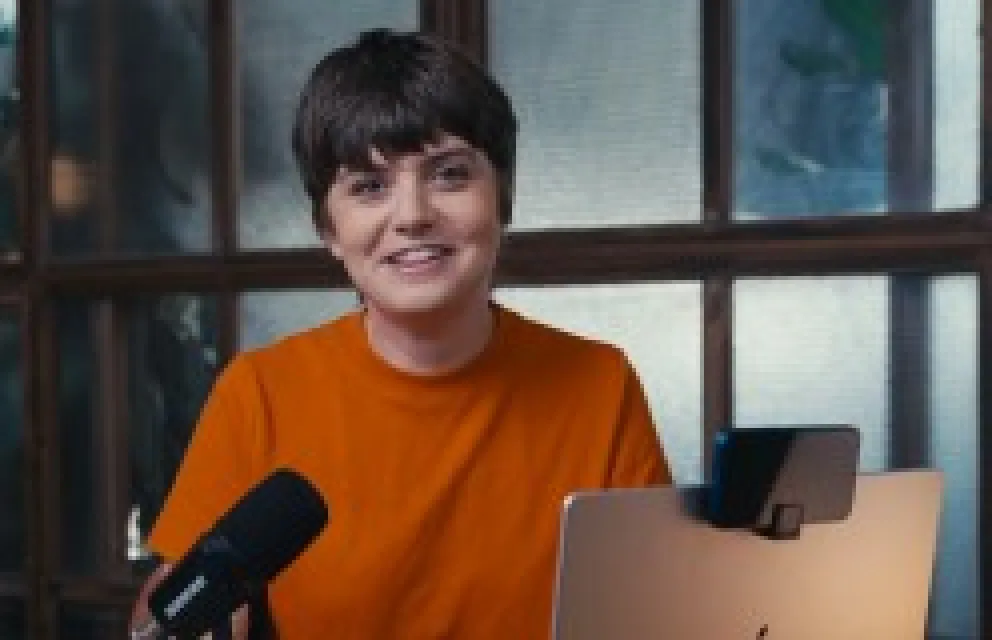 Person with short dark hair smiling, wearing an orange shirt, sitting at a desk with a microphone and a laptop.